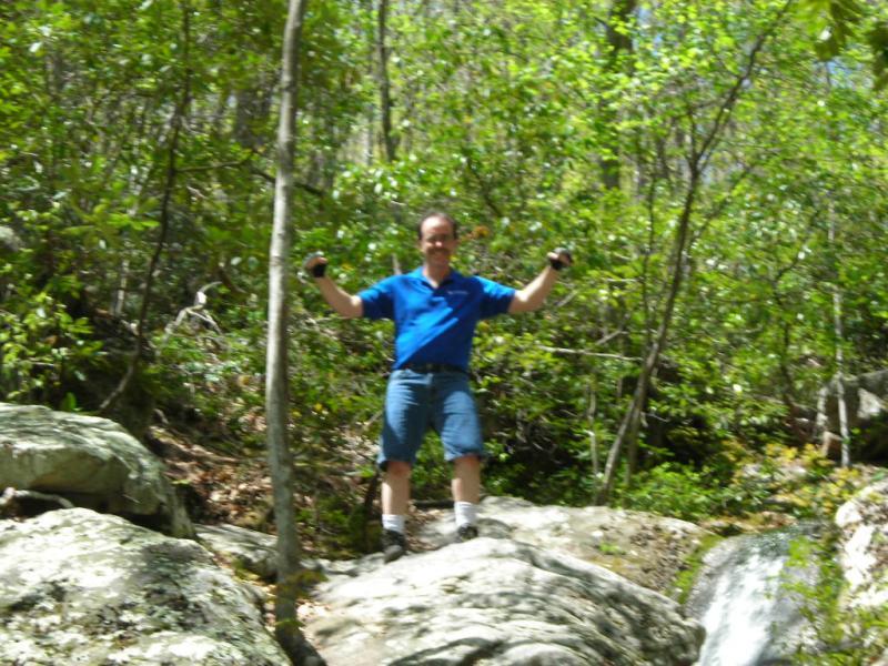 A person wearing a blue shirt and shorts stands on a rock in a lush, green forest, raising their arms with a smile. Sunlight filters through the trees, highlighting the natural surroundings. Douthat State Park mountain bike trail.