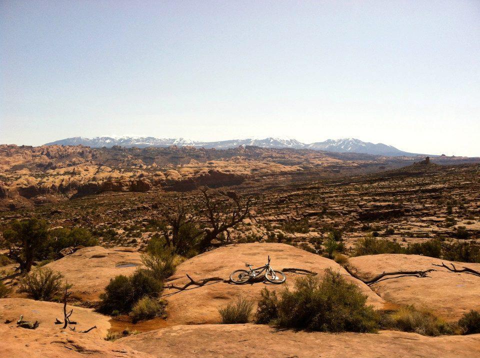 A scenic view of a rocky landscape with distant snow-capped mountains under a clear blue sky. In the foreground, a bicycle rests on a large rock, surrounded by desert vegetation and an expansive natural vista. Captain Ahab mountain bike trail.