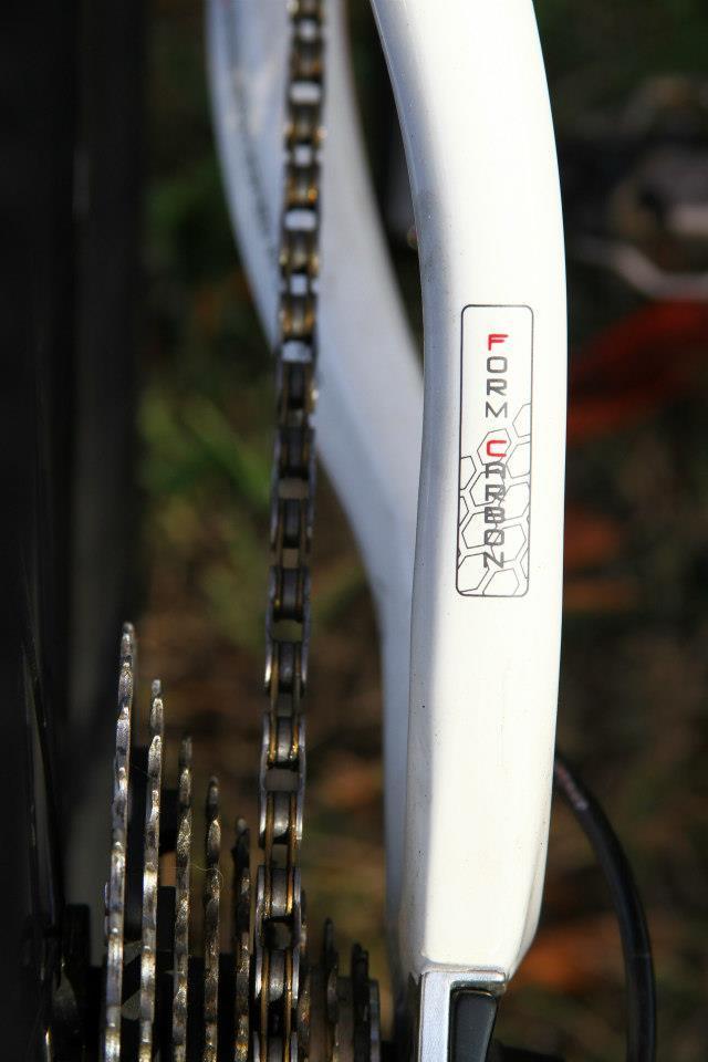 Rocky Mountain Vertex 50: Close-up view of a bicycle's rear derailleur and chain, featuring a white carbon frame with the text "FORM CARBON" displayed on the side. The chain is partially visible, showcasing the gears and links, set against a blurred natural background.
