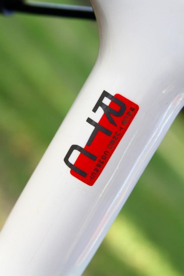 Rocky Mountain Vertex 50: A close-up of a white bicycle frame showcasing a logo with red and black elements, featuring stylized letters and numbers. The background is softly blurred green grass, suggesting an outdoor setting.