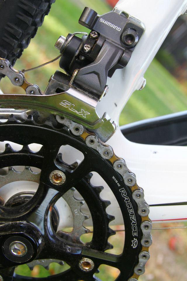 Rocky Mountain Vertex 50: Close-up view of a bicycle's drivetrain, featuring a Shimano SLX derailleur and a black Race Face chainring. The image highlights the intricate details of the components, set against a blurred green background.