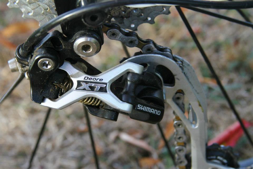 Rocky Mountain Vertex 50: Close-up image of a Shimano Deore XT bicycle rear derailleur, showcasing its silver and black components, attached to a chain and gear system against a natural background.