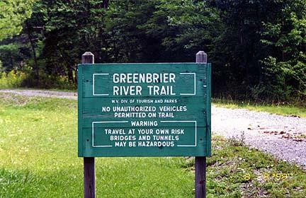 A green sign marking the Greenbrier River Trail, stating that no unauthorized vehicles are permitted and cautioning that travel is at your own risk due to potentially hazardous bridges and tunnels. The sign is surrounded by greenery and is positioned along a gravel path. Greenbrier River Trail mountain bike trail.