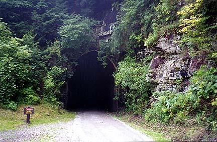 A dark, arched tunnel entrance surrounded by lush green foliage and rocky cliffs, with a gravel pathway leading toward it. A small sign is visible on the left side of the path near the tunnel entrance. Greenbrier River Trail mountain bike trail.