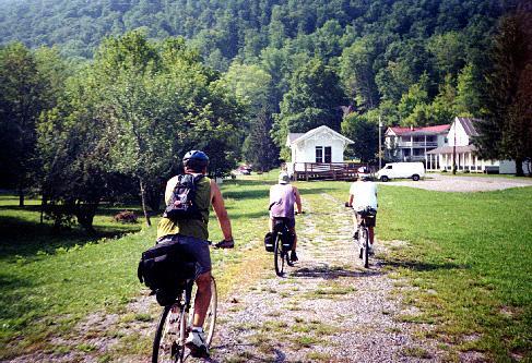 Three cyclists ride along a gravel path in a green landscape, surrounded by trees and hills. In the background, a white house and a small building are visible, along with a parked van. The scene conveys a sense of outdoor adventure and tranquility. Greenbrier River Trail mountain bike trail.