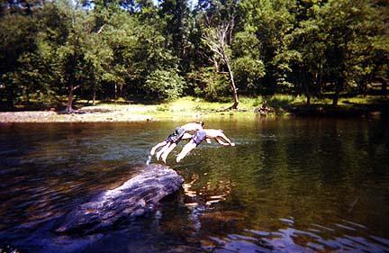 A person jumping off a large rock into a clear, calm river surrounded by lush green trees. The scene captures a moment of outdoor adventure and summer fun. Greenbrier River Trail mountain bike trail.