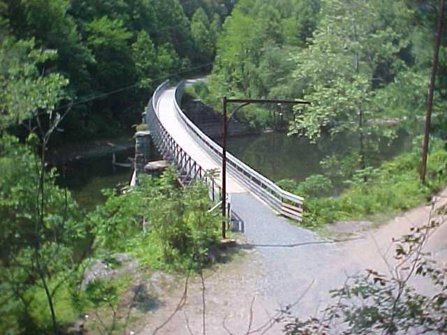 A narrow wooden bridge arching over a peaceful river, surrounded by lush greenery and trees. The path leading to the bridge is gravel, with a power pole on the right side of the image. The scene conveys a tranquil, natural setting ideal for outdoor activities. Greenbrier River Trail mountain bike trail.