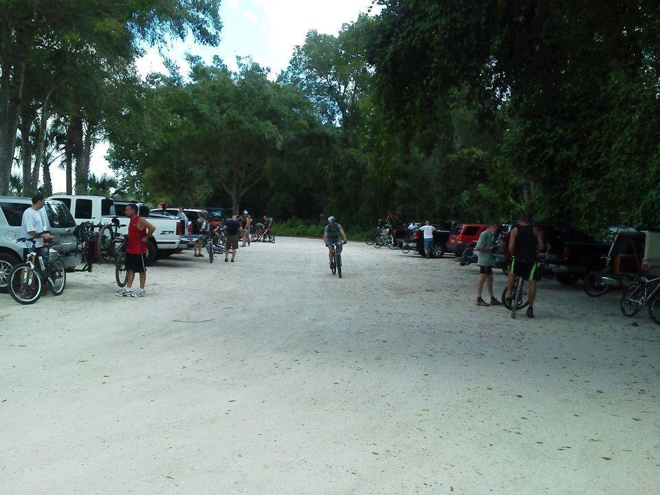 A dirt parking area surrounded by trees, filled with several parked vehicles. People are seen preparing their bicycles, some standing beside their bikes while others are getting ready to ride. The scene captures an outdoor recreational atmosphere. Chuck Lennon Park mountain bike trail.
