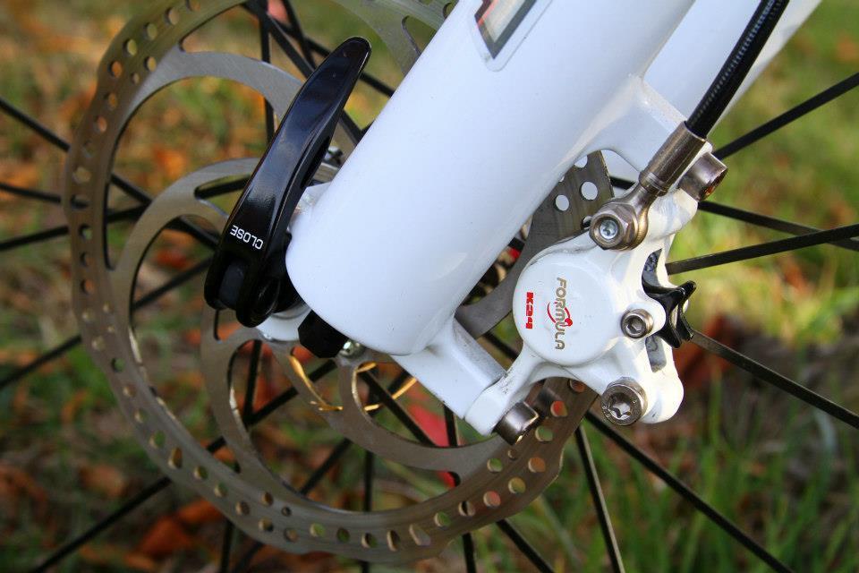 Rocky Mountain Vertex 50: Close-up view of a bike's disc brake system, featuring a white brake caliper and rotor, with a black lever labeled "CLOSE." The background includes blurred green grass and orange leaves, indicating an outdoor setting.