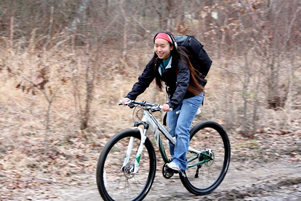 Specialized Myka Disc: A person riding a mountain bike along a dirt trail, wearing a black jacket and jeans, with long hair flowing behind them. They have a backpack and are smiling as they navigate the outdoor path, surrounded by sparse trees and dry grass.