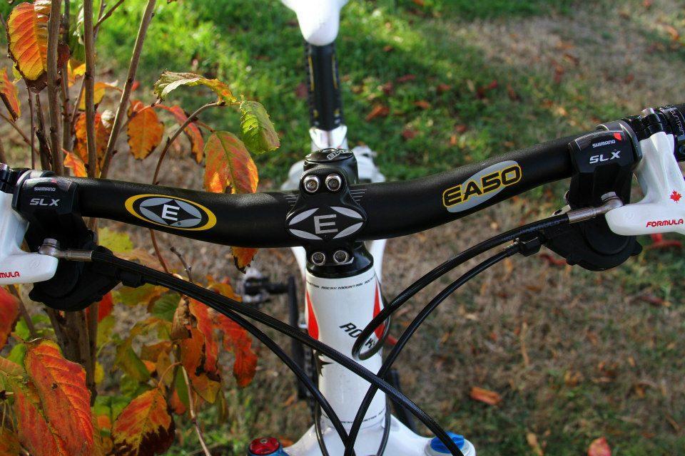 Rocky Mountain Vertex 50: Close-up view of a bicycle's handlebars, featuring technical components such as the stem and brake levers. The handlebar is black with logos, including "E" and "EA50," and there are visible cables leading to the brakes. Surrounding the handlebars are colorful autumn leaves, set against a grassy background.