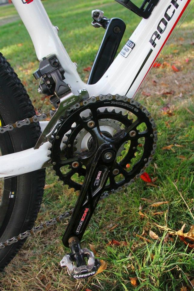 Rocky Mountain Vertex 50: Close-up view of a bicycle's crankset and chainring, featuring a black Shimano pedal and a silver chain with a white frame background. The image is set on grass with a few fallen leaves, showcasing a detailed angle of the bike's drivetrain components.