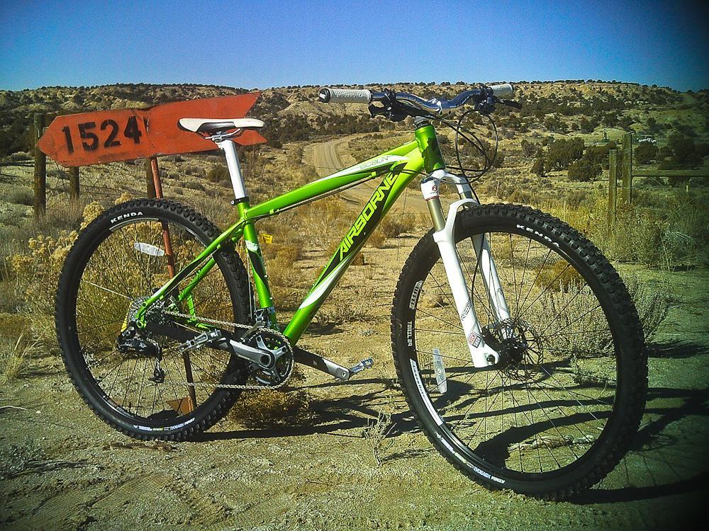 Airborne Goblin: A green mountain bike with white front forks is positioned on a dirt path, with the background featuring dry, desert-like terrain and a clear blue sky. An old wooden sign with the number "1524" can be seen nearby, adding to the rustic outdoor setting.