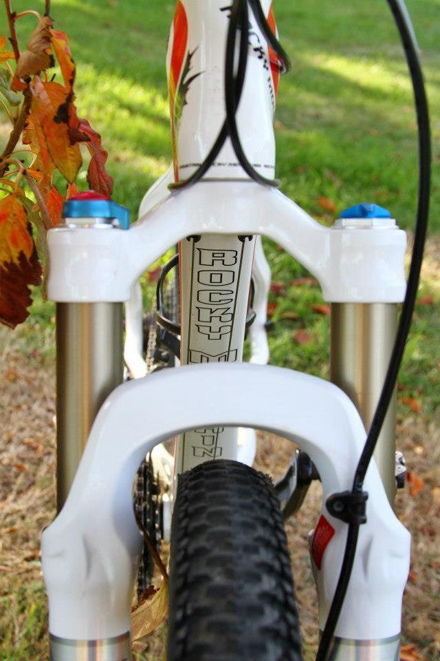 Rocky Mountain Vertex 50: Close-up view of the front suspension of a mountain bike, featuring a white fork and a textured tire, with colorful autumn leaves in the background and green grass underneath. The bike's frame and components are partially visible.