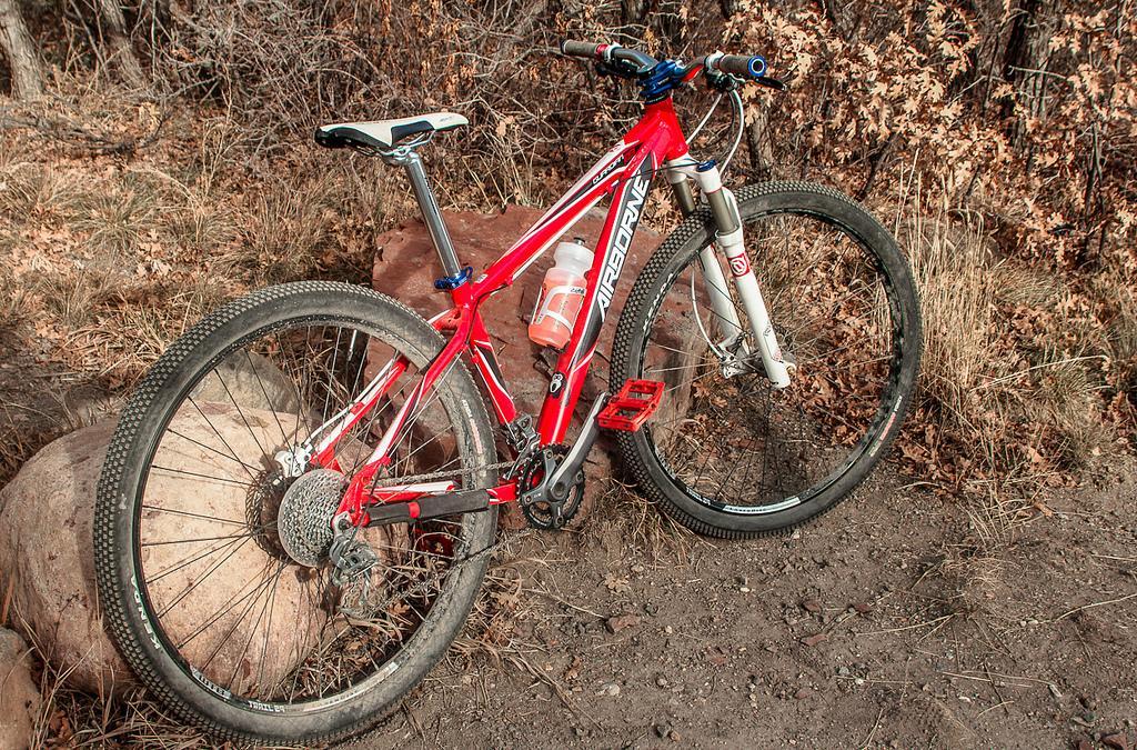Airborne Guardian: A red mountain bike leaning against a large rock on a dirt trail, surrounded by dry grass and sparse bushes. The bike features a suspension fork, wide tires, and a water bottle attached to the frame. Sunlight casts soft shadows, highlighting the bike's design and the natural surroundings.