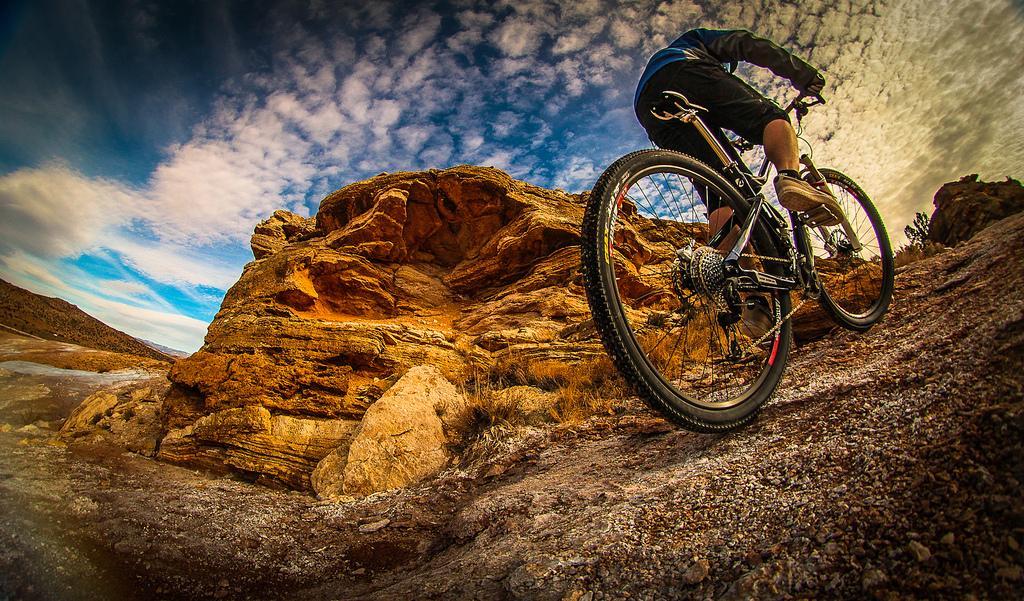 A mountain biker ascends a rocky trail, showcasing the bike's wheel and the rider's lower body. The backdrop features dramatic rock formations and a colorful sky with clouds. The terrain is rugged, indicating an adventurous outdoor setting. White Ridge Bike Trails mountain bike trail.