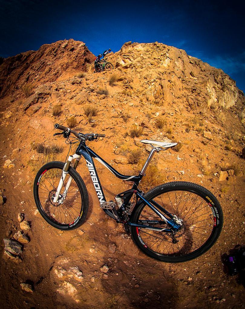 A mountain bike rests on rocky terrain, with a rider navigating a steep, rugged slope in the background. The scene is set against a blue sky, showcasing the challenging landscape of a mountain biking trail. White Ridge Bike Trails mountain bike trail.