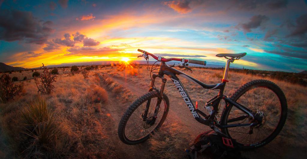 Airborne Hobgoblin: A mountain bike rests on a dirt trail against the backdrop of a vibrant sunset, with colorful clouds illuminating the sky. The surrounding landscape features dry grasses and shrubs, creating a serene outdoor atmosphere perfect for biking adventures.