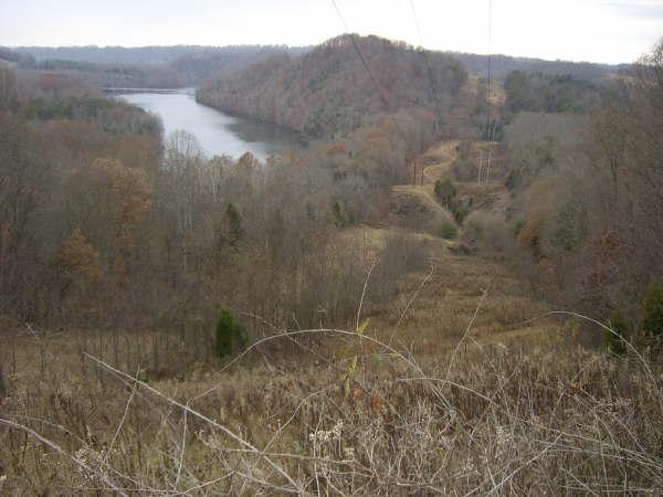 A scenic view of a tranquil river surrounded by wooded hills and bare trees, with a foreground of overgrown grass and twigs. The landscape is captured on a cloudy day, suggesting a peaceful, natural environment in late autumn or early winter. Warriors' Path State Park mountain bike trail.