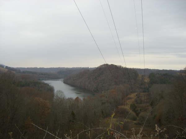 A scenic view of a river winding through a valley surrounded by wooded hills, under an overcast sky with power lines running overhead. Warriors' Path State Park mountain bike trail.