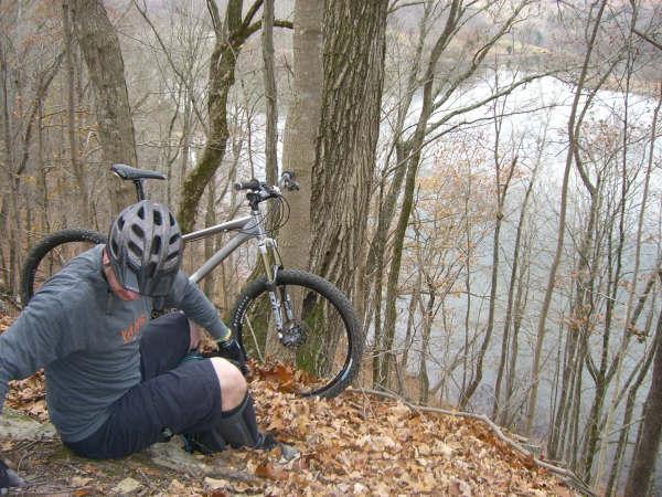 A mountain biker in a helmet sitting on a rocky slope covered with fallen leaves, with a bicycle resting beside him. In the background, trees line the slope with a view of a river flowing below. The scene captures a moment of pause in a natural outdoor setting. Warriors' Path State Park mountain bike trail.