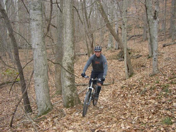 A person riding a mountain bike on a wooded trail, surrounded by trees and autumn leaves. The cyclist is wearing a helmet and athletic gear, navigating a dirt path in a natural setting. Warriors' Path State Park mountain bike trail.
