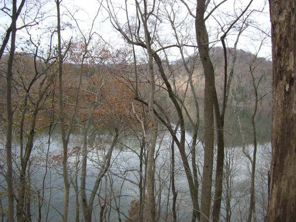 A serene landscape featuring a calm lake surrounded by bare trees and a rocky hillside in the background. The scene captures a sense of tranquility with soft, muted colors, indicating late autumn or early winter. Warriors' Path State Park mountain bike trail.