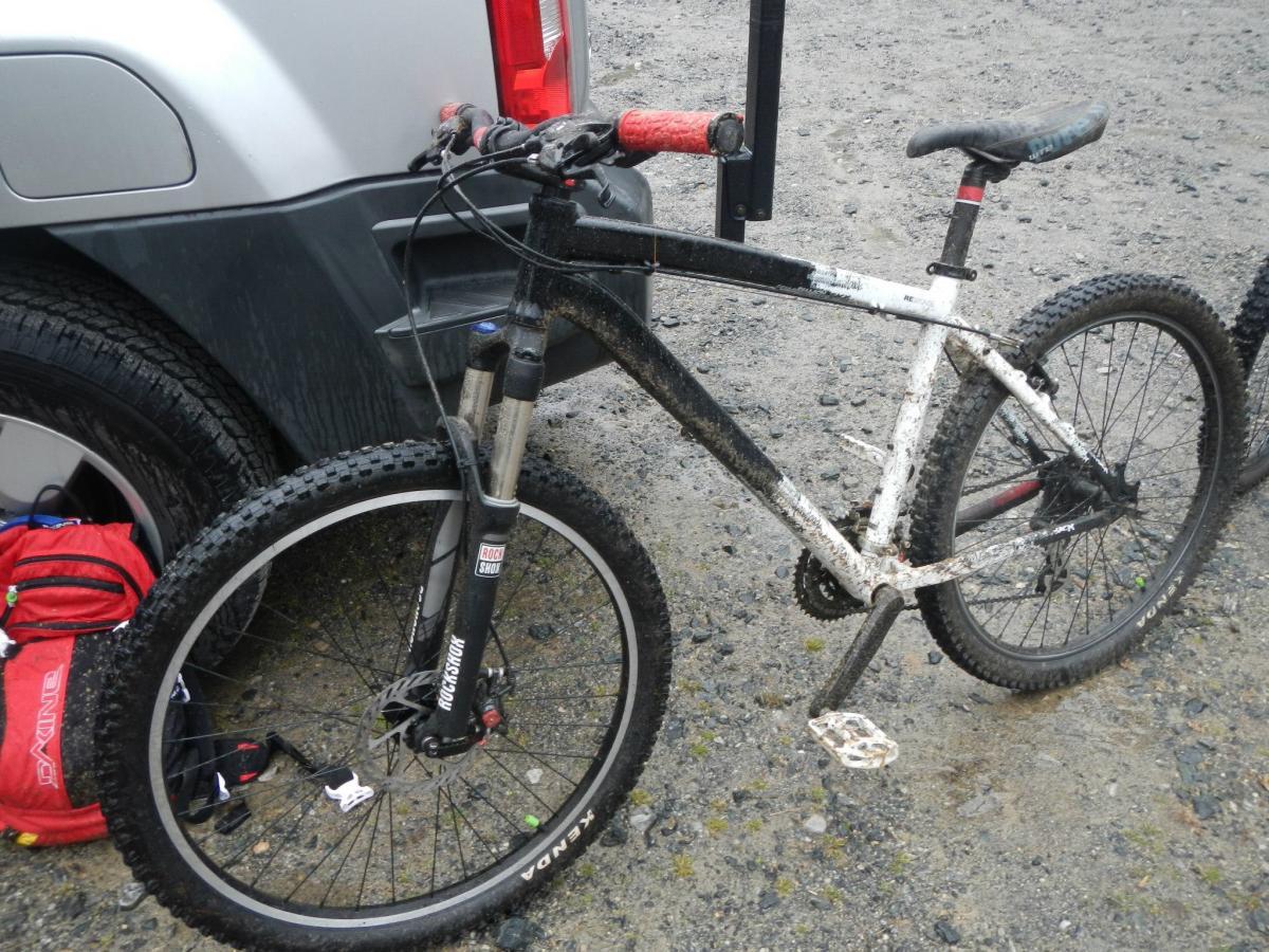 Diamondback Response: A mountain bike with a black and white frame, covered in mud, parked next to a vehicle. The bike has thick, knobby tires and a visible suspension system. A red backpack is partially visible on the ground nearby, against a gravel background.