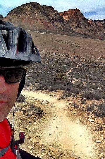 A person wearing a helmet and sunglasses poses for a selfie on a mountain biking trail. In the background, a rugged mountainous landscape is visible, with a winding dirt path leading away. The terrain appears arid and rocky, suggesting a remote outdoor setting. Cottonwood Valley North mountain bike trail.