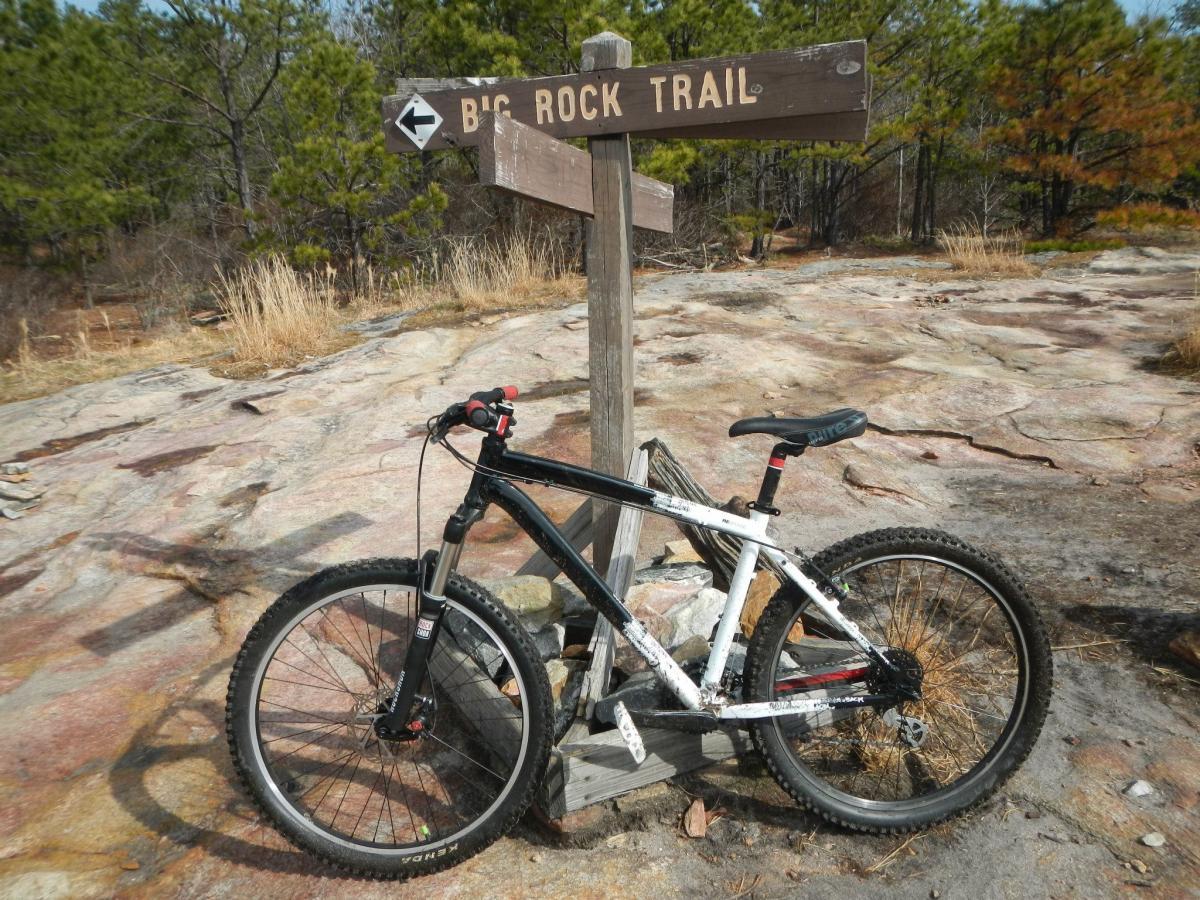 Diamondback Response: A mountain bike leaning against a wooden trail sign labeled "Big Rock Trail" in a natural outdoor setting. The background features rocky terrain and sparse pine trees under a clear sky.