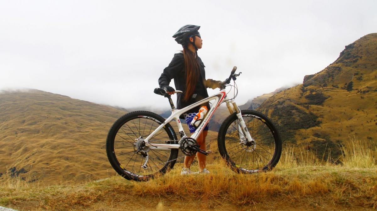 Rocky Mountain Vertex 50: A person wearing a helmet and a black jacket stands on a grassy hillside, holding a mountain bike. Behind them, a landscape of rolling hills covered in brown grass meets a cloudy sky. The scene captures a sense of adventure and the beauty of the outdoors.