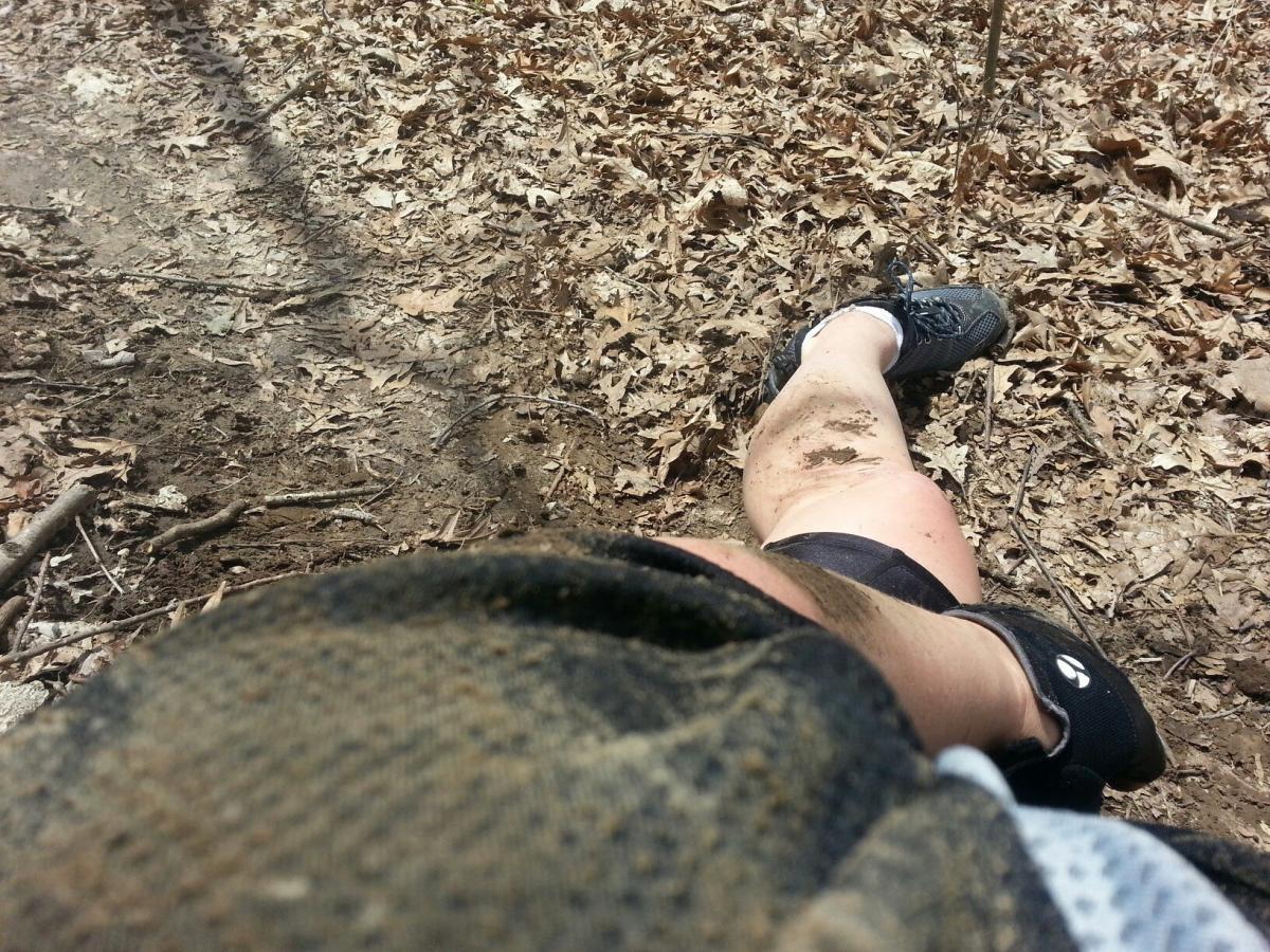 A person's leg is visible, covered in dirt and mud, while resting against the ground. The surrounding area is filled with dry leaves and twigs, indicating an outdoor setting, possibly a hiking trail. The person is wearing black athletic shoes and dark shorts, and the perspective suggests they are sitting or lying down. Illiniwek Forest Preserve mountain bike trail.