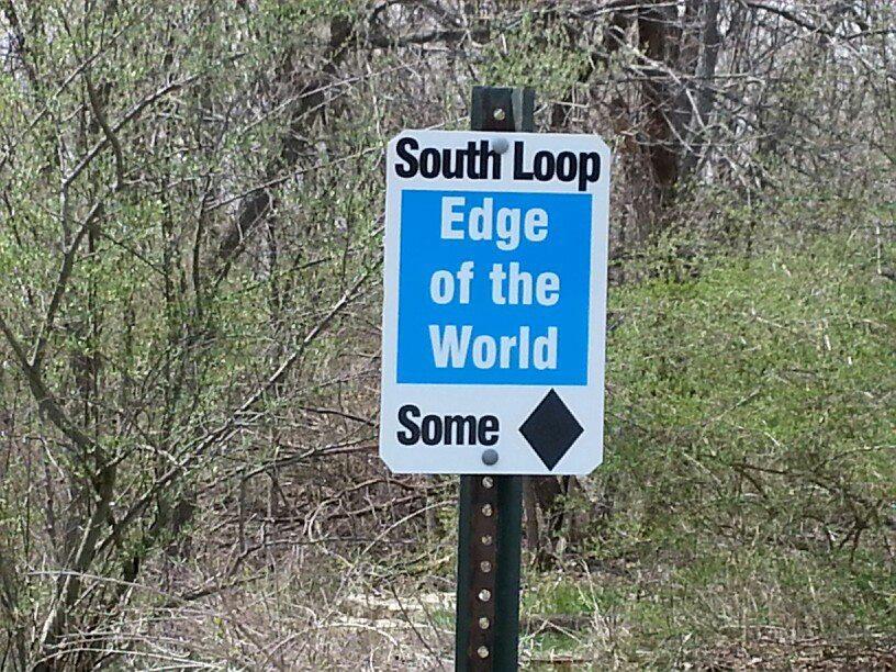 A sign indicating "South Loop Edge of the World" displayed against a backdrop of trees and greenery. The sign has a blue and white color scheme with the word "Some" at the bottom, accompanied by a diamond shape. Wildlife Prairie Park mountain bike trail.