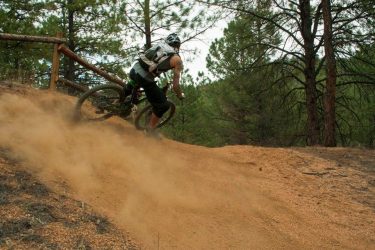 A mountain biker navigating a dirt trail, kicking up dust as they accelerate around a turn in a forested area. Pine trees surround the path, creating a natural backdrop for the action. Buffalo Creek mountain bike trail.