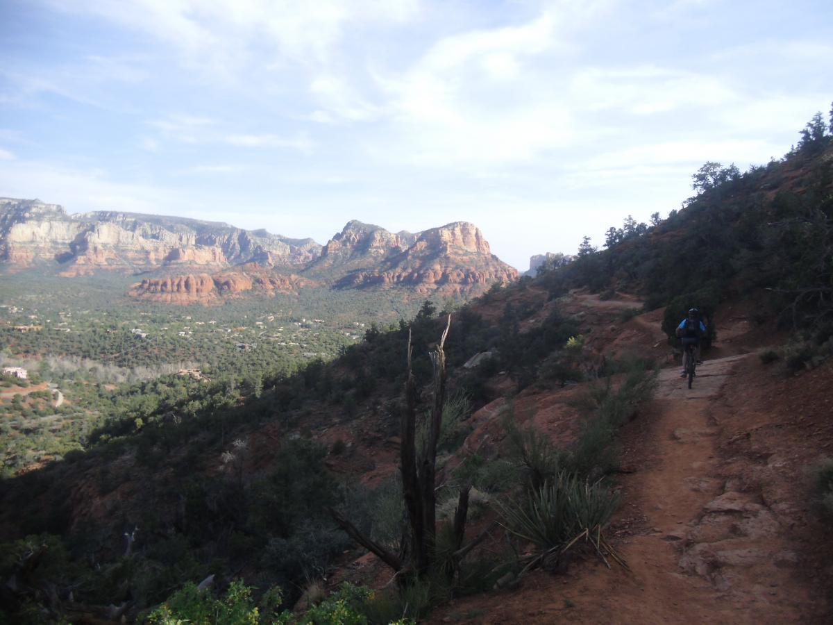 A cyclist riding along a rocky trail with red rock formations in the background, surrounded by greenery and a clear blue sky. Airport Mesa Trail Network mountain bike trail.