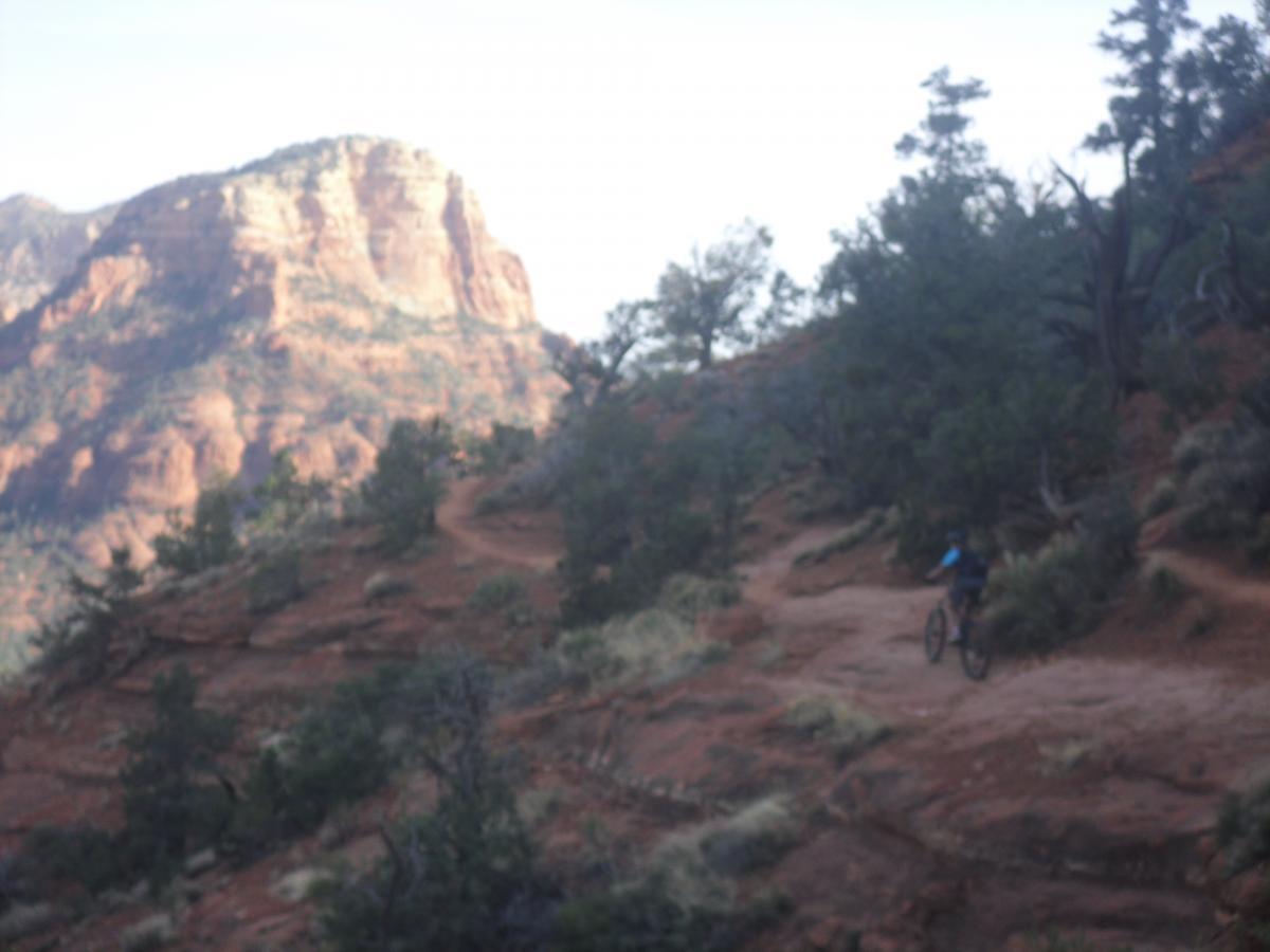 A mountain biker riding along a winding dirt trail through red rock terrain, surrounded by sparse vegetation and distant mountains under a clear sky. Airport Mesa Trail Network mountain bike trail.