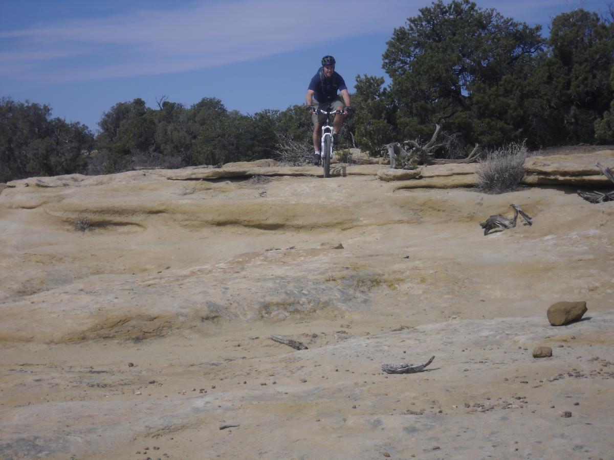 A person riding a mountain bike on rocky terrain, with shrubs and trees in the background. The sky is partly cloudy, suggesting a sunny day. Alien Run Trail mountain bike trail.