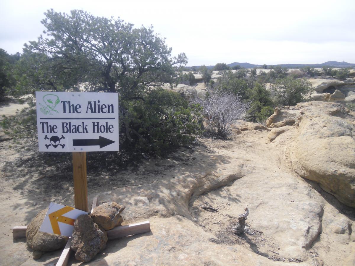A signpost in a rocky desert landscape, indicating directions to "The Alien" and "The Black Hole," with a graphic of an alien and hazard symbols. The scene features sparse vegetation and a cloudy sky. Alien Run Trail mountain bike trail.