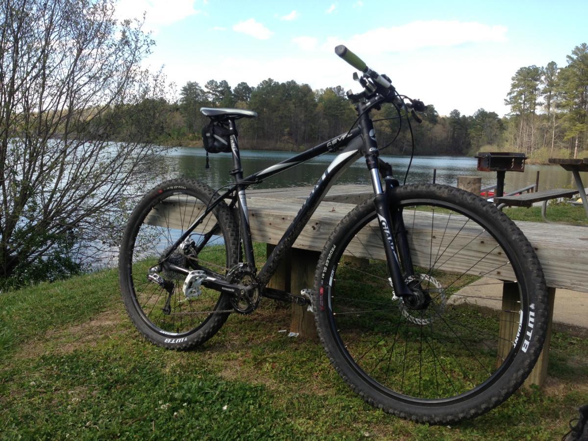 Giant 29er Revel 0: Mountain bike resting on a wooden platform by a serene lake, surrounded by lush greenery and trees. The bike features thick tires and a sleek frame, with the water reflecting the sky and nature in the background.