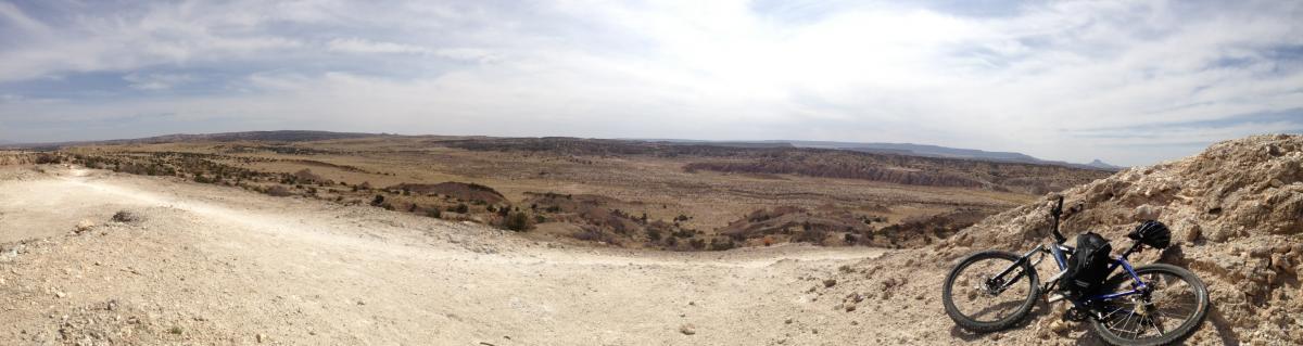 Specialized Hardrock Pro Disc: Panoramic view of a vast landscape with rolling hills and a distant horizon, featuring a dirt path in the foreground and a mountain bike resting on the ground with a helmet nearby. The sky is partly cloudy, creating a serene outdoor scene.