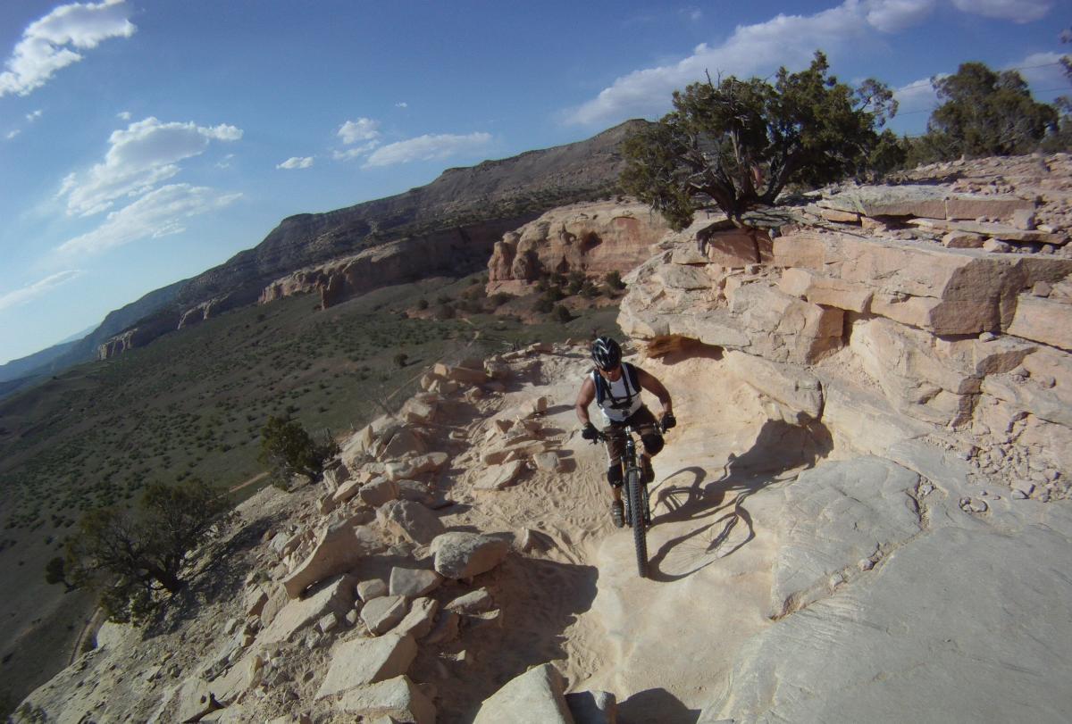 A mountain biker navigating a rocky trail on a sunny day, surrounded by rugged terrain and distant hills under a blue sky with clouds. Mary's Loop / Horsethief Bench mountain bike trail.