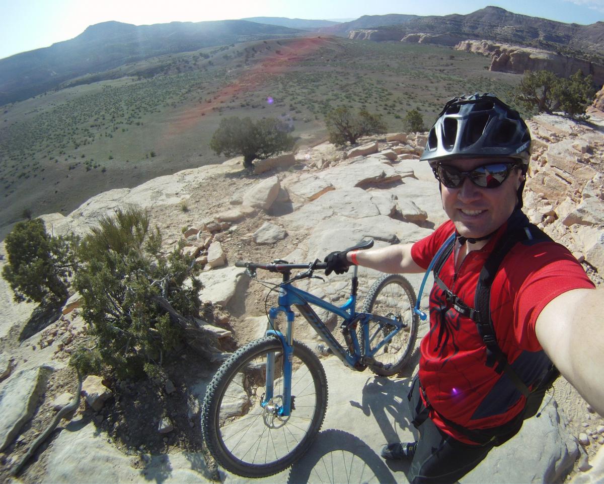A mountain biker wearing a red shirt and sunglasses takes a selfie while standing with his blue mountain bike on a rocky outcrop. In the background, a vast landscape of green hills and distant mountains is visible under a clear sky. Mary's Loop / Horsethief Bench mountain bike trail.
