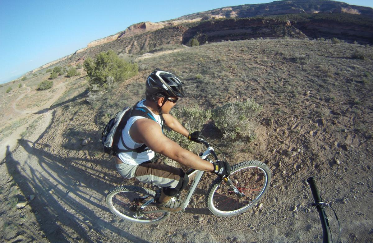 A person riding a mountain bike on a dirt trail surrounded by rocky terrain and sparse vegetation, with a clear blue sky in the background. The cyclist is wearing a helmet and a backpack, focused on the ride. Rustlers Loop mountain bike trail.