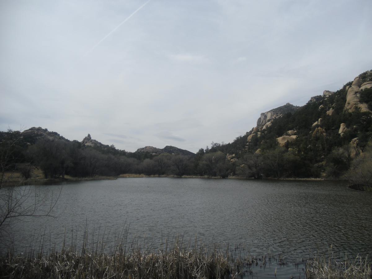 A serene landscape featuring a calm lake surrounded by rugged hills and rocky formations. The sky is overcast, with soft clouds creating a tranquil atmosphere. Sparse trees line the shore, and tall grasses gently brush the water's edge. Granite Basin Loops mountain bike trail.