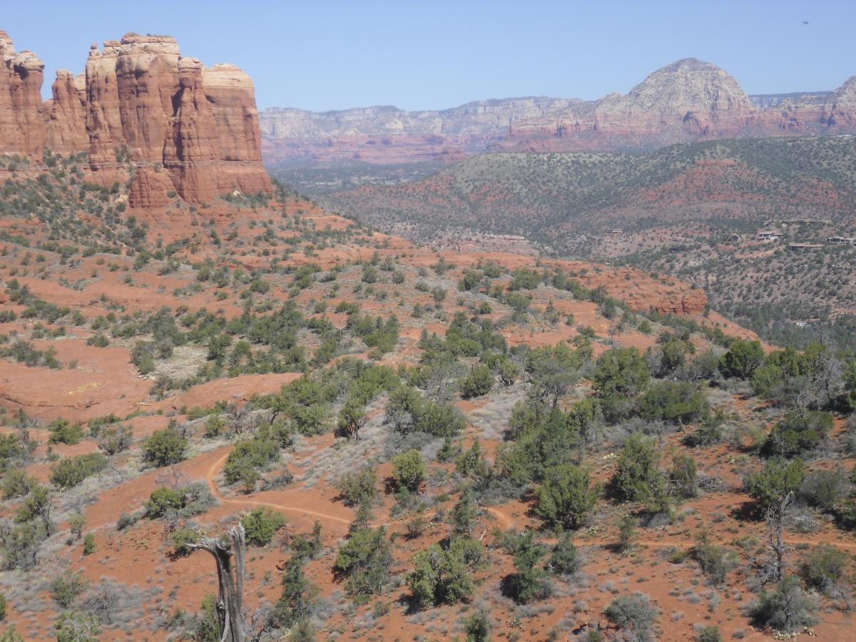 A panoramic view of a rugged desert landscape featuring red sandstone formations, green vegetation, and distant mountains under a clear blue sky. A winding dirt path can be seen through the terrain, showcasing the natural beauty of the area. Hiline mountain bike trail.