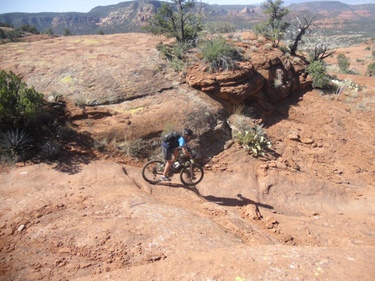 A person riding a mountain bike over rocky terrain in a desert landscape, surrounded by sparse vegetation and red rock formations, with distant hills and blue sky in the background. Hiline mountain bike trail.