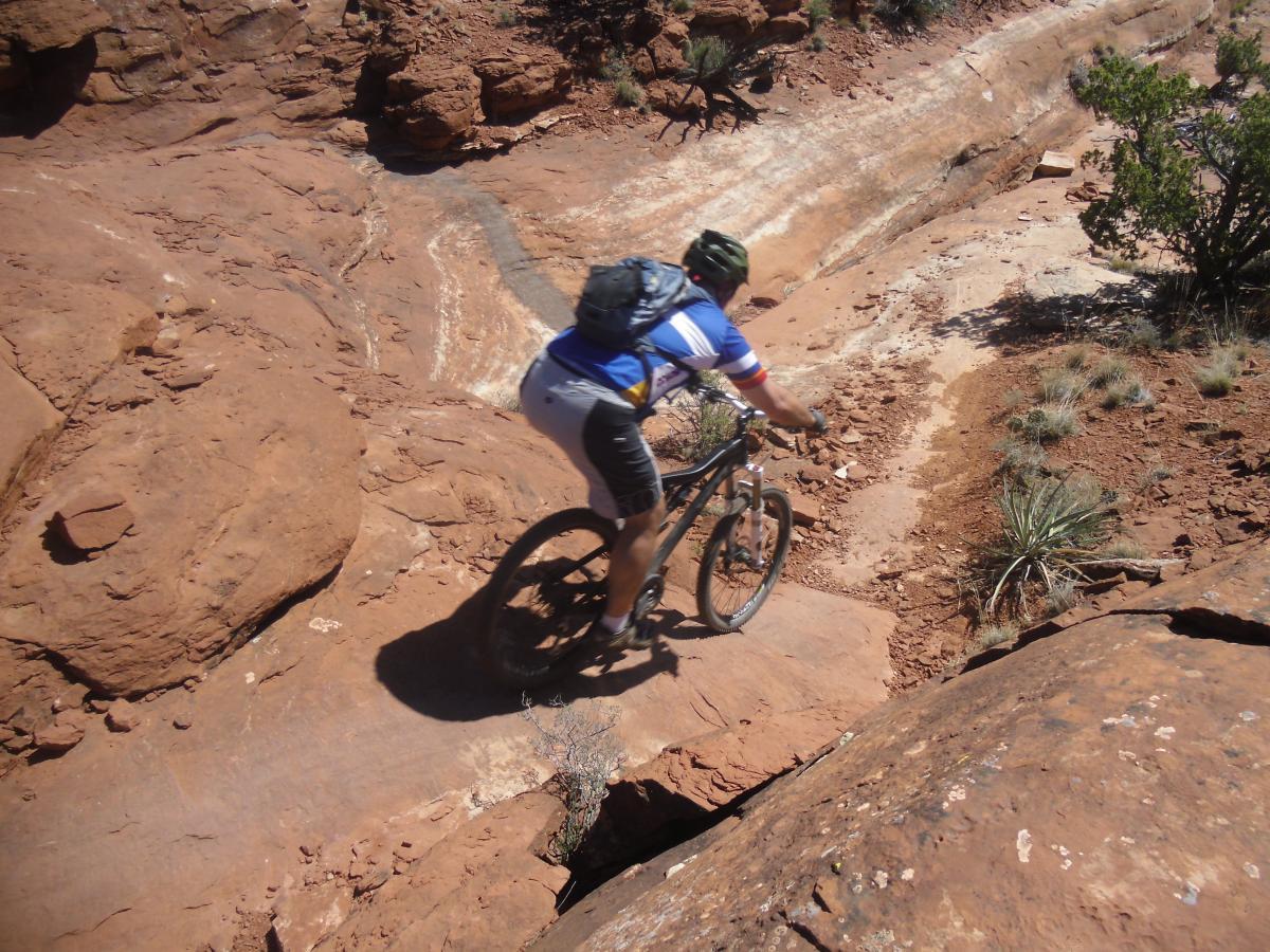 A mountain biker navigating a rocky trail, surrounded by red earth and sparse greenery, as he descends a steep incline. The terrain features rough, uneven rocks and a winding path, highlighting the challenging conditions of off-road cycling. Hiline mountain bike trail.