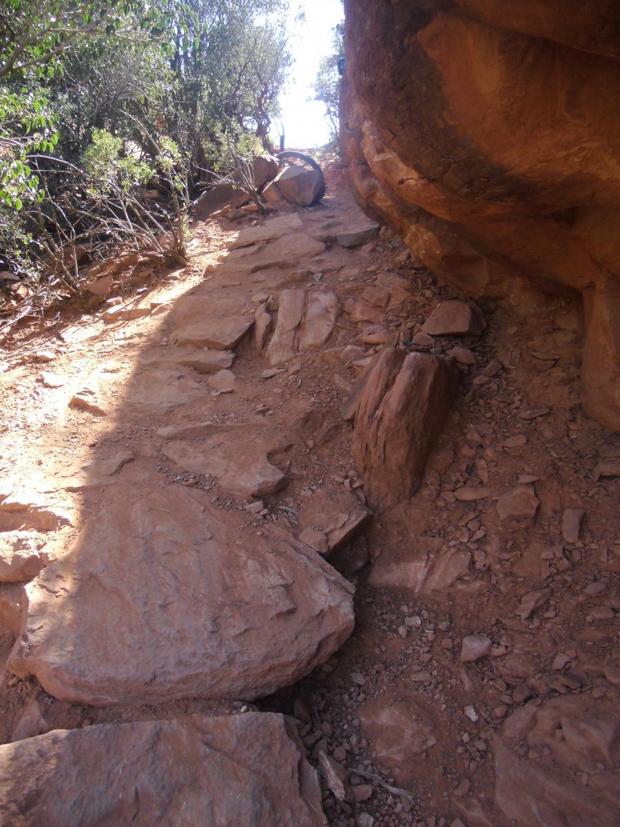 A rocky trail winding through a sunlit landscape, flanked by rugged cliffs and sparse vegetation. The path is uneven, with large stones and dirt, leading upwards into the distance. Hiline mountain bike trail.