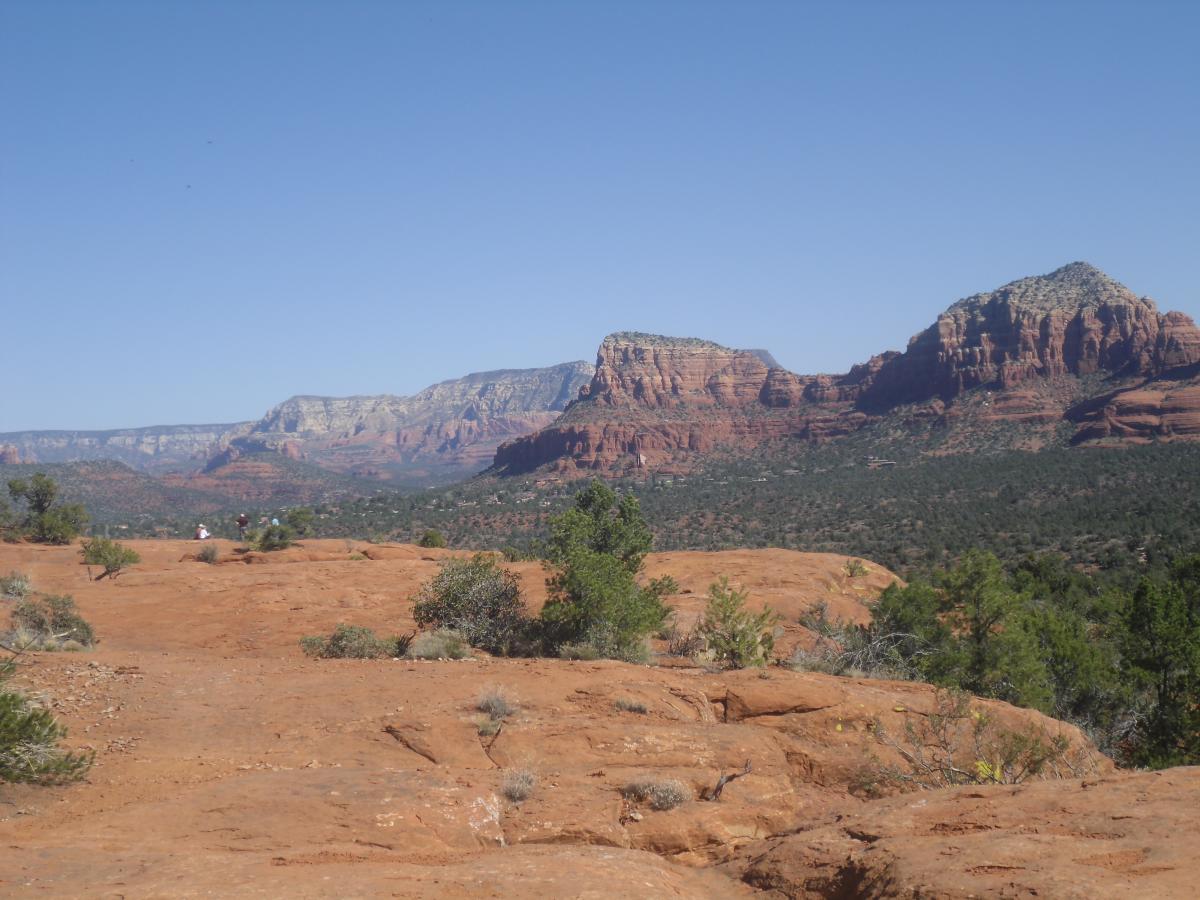 Scenic view of red rock formations and distant mountains under a clear blue sky, with minimal vegetation in the foreground and a few people in the distance. Hiline mountain bike trail.