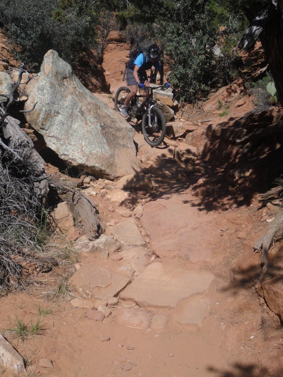 A mountain biker navigating a rocky trail with a large boulder on one side, surrounded by vegetation and sandy terrain. The cyclist is focused on the path ahead, showcasing the challenge of the off-road environment. Hiline mountain bike trail.